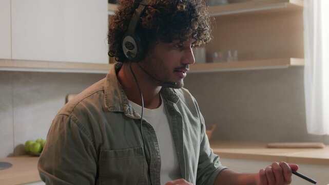 Hispanic Indian Arabian man in headphones teaching online at home kitchen laptop video call ethnic male coach psychologist remote e-learning lesson writing notes virtual conference education training