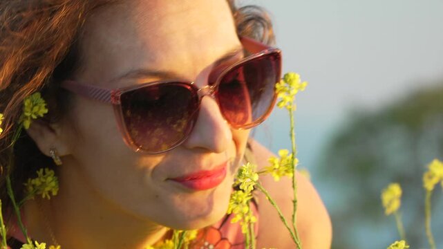 Smiling young woman in sunglasses enjoys sniffing yellow wildflowers outdoors during a golden hour sunset. Close-up zoom shot.