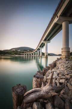 View of the bridge over Lago di Cingoli with calm turquoise water, rocky shoreline, and wooden posts under a clear sky at sunset in Cingoli, Marche, Italy.