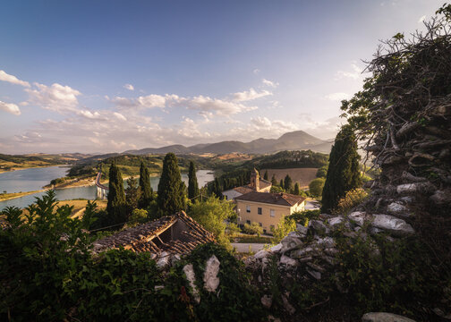 View of Lago di Cingoli surrounded by cypress trees, rolling hills, and historic architecture under a clear blue sky during golden hour in Cingoli, Marche, Italy.