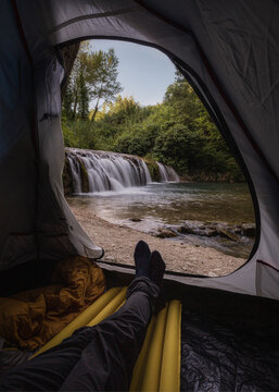 View of Lago di Cingoli with a waterfall flowing into a stream, seen from inside a camping tent with a person's legs resting on a sleeping pad in Cingoli, Marche, Italy.