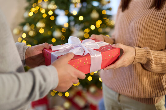 Christmas couple exchange gift present box at home. Hands share a red ribboned box before a glowing festive tree, warm lights and bokeh shaping cozy holiday mood. New Yar celebration and giving.