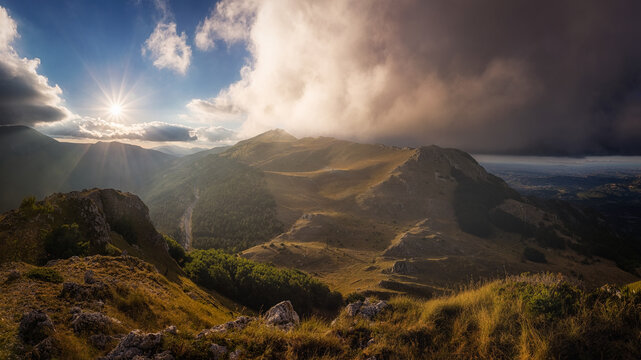 View of the Sibillini Mountains with dramatic clouds, sunburst, and grassy slopes, Bolognola, Marche, Italy.