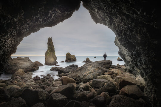View of a person standing on the rocky shore of Valahnukamol looking out at sea stacks from inside a sea cave in Reykjanesbaer, Iceland.