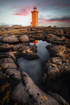 View of Stafnesviti lighthouse standing on a rugged volcanic coastline with a reflection in a tidal pool under a dramatic sunset sky in Sudurnesjabaer, Iceland.