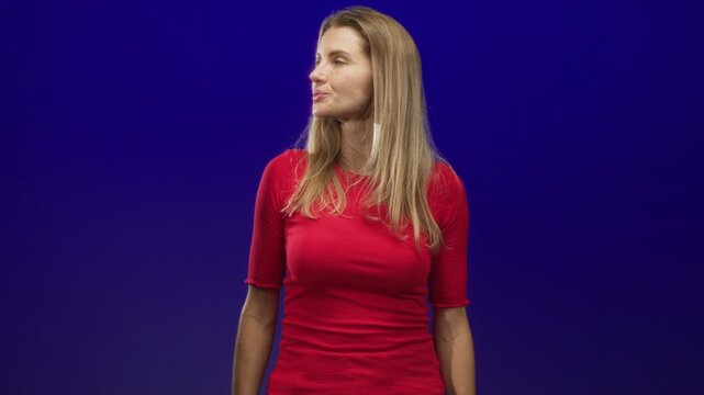 Woman turns head left wearing a red top, standing with relaxed arms against a deep blue studio wall under soft directional light; contemplation.
