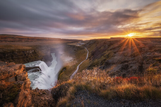 View of Gullfoss Falls at sunset with dramatic golden light illuminating the rugged canyon, rocky cliffs, and a winding walking path in Hrunamannahreppur, Iceland.