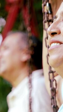 Indian Asian mid age couple in their fifties with gray hair sitting on swings in the park, smiling at the camera, enjoying a peaceful moment outdoors in daylight