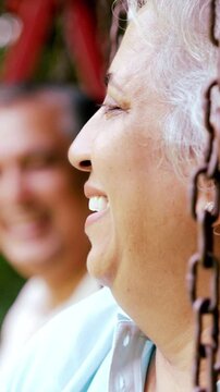 Indian Asian mid age couple in their fifties with gray hair sitting on swings in the park, smiling at the camera, enjoying a peaceful moment outdoors in daylight