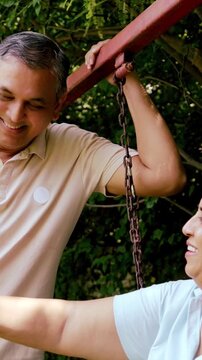 Indian Asian mid age couple in their fifties with gray hair sitting on swings in the park, smiling at the camera, enjoying a peaceful moment outdoors in daylight