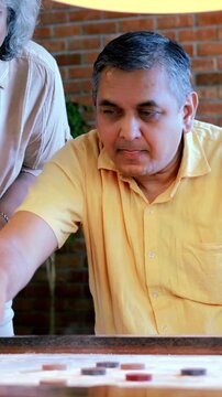 Indian Asian mid age couple in their fifties with gray hair playing carrom, enjoying an indoor board game with a light and happy mood
