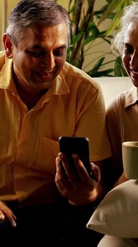 Indian Asian couple in their fifties with gray hair enjoying tea or coffee by a sunny window, relaxing on the sofa and spending quality time together in the morning