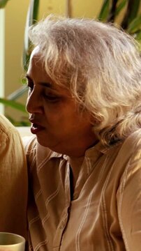 Indian Asian couple in their fifties with gray hair making a video call on laptop or smartphone, sitting on a sofa near a sunlit window at home, enjoying the moment