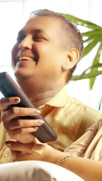 Indian Asian couple in their fifties with gray hair making a video call on laptop or smartphone, sitting on a sofa near a sunlit window at home, enjoying the moment