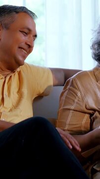 Indian Asian couple in their fifties with grey hair sitting cozily on a sofa, laughing heartily together, enjoying an funny moment in the warm evening light