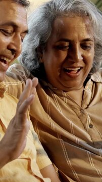 Indian Asian couple in their fifties with gray hair making a video call on laptop or smartphone, sitting on a sofa near a sunlit window at home, enjoying the moment