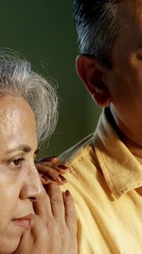 Indian Asian couple in their fifties with grey hair standing close by a large window, bathed in morning sunlight, both gazing outside with serious expressions, reflecting in the moment