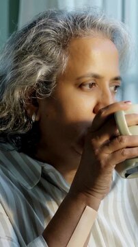 Indian Asian couple in their fifties with grey hair standing by a sunlit window, holding coffee or tea mugs, sharing a moment of quiet with pleasant expressions