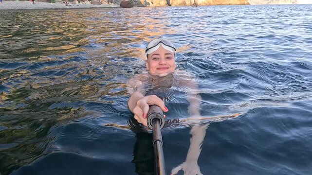 Swimming tourist recording self-video with monopod while floating in calm sea near rocky shore. Action camera tracking view.