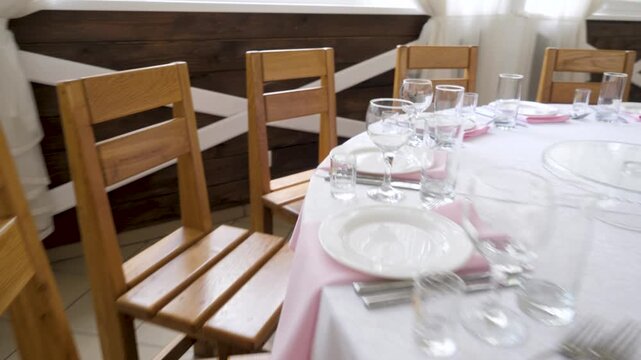 A round banquet table decorated with white tablecloth, pink napkins, stemware, plates, and floral centerpieces stands surrounded by wooden chairs inside a venue.