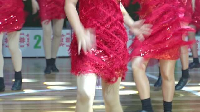 A troupe of female dancers in matching red fringe dresses perform synchronized dance moves on a wooden stage.