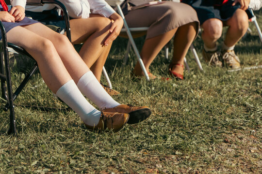 A row of people sit on portable folding chairs outdoors on sunny green grass wearing skirts, socks, and vintage shoes.