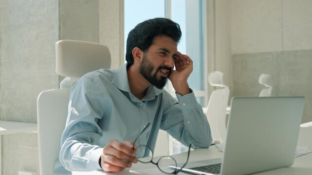 Tired Arabian Indian business man rubbing temple removing glasses at laptop office male businessman head cramp painful ache tension migraine sick ethnic worker fatigue eyestrain computer desk stress