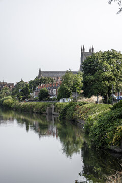 View of Worcester Cathedral rising above the River Severn with lush riverbanks, historic buildings, and calm water reflecting the scene in Worcester, England, United Kingdom.