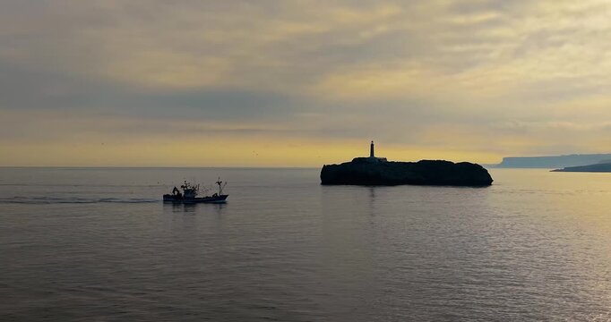 Cinematic aerial view of a traditional fishing boat entering Santander bay at golden sunrise, passing Mouro Island lighthouse with a catch of anchovies, mackerel, and hake in Cantabria, Northern Spain