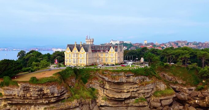 Panoramic aerial flyover of the iconic royal Magdalena Palace (Palacio de la Magdalena) on a rocky peninsula in Spain, with Santander city visible in the background.