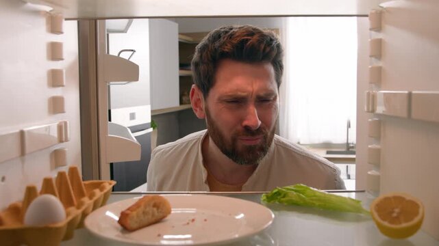 Confused Caucasian middle aged man sniffing bad smell spoiled food POV inside empty refrigerator with egg lettuce lemon bread male guy checking rotten leftovers odor stench unpleasant kitchen problem