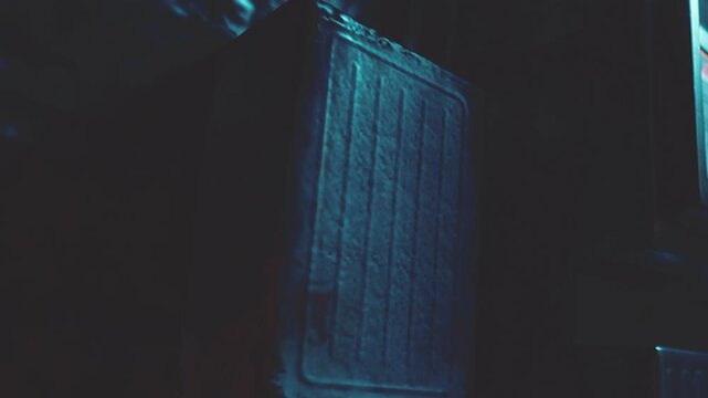 A small room is dimly lit with shadows. A wooden crate sits against a wall. The room shows signs of night with limited light coming from a window.