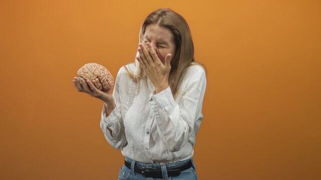 Woman holding brain in left hand, right hand covering mouth and nose against orange studio backdrop; anxiety reflection.