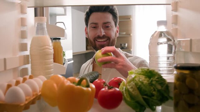 Hungry smiling Caucasian man choosing healthy vegetables eating apple in kitchen male searching food deciding meal chef enjoying fresh fruit health dish diet nutrition delivery POV inside refrigerator