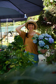 Asian Woman Smiling at Outdoor Flower Market Holding Blue Hydrangeas in Straw Hat