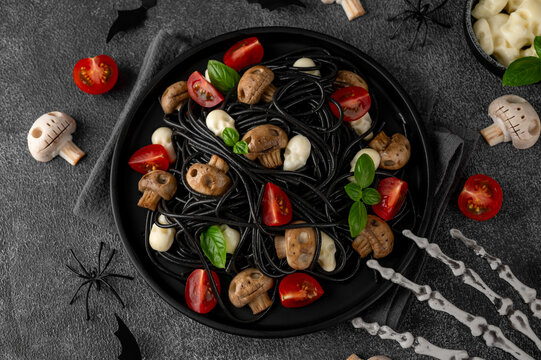Halloween-themed food. Black squid ink spaghetti with mozzarella skulls, mushrooms, cherry tomatoes and fresh basil. Creative spooky food styling for festive autumn celebrations.