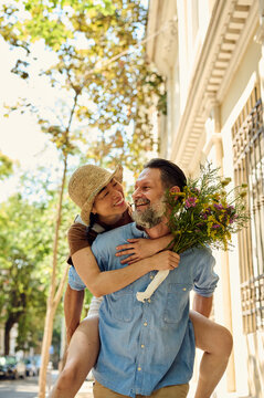 Happy Couple Piggyback On Urban Street With Bouquet &mdash; Asian Woman And Bearded Man Laughing