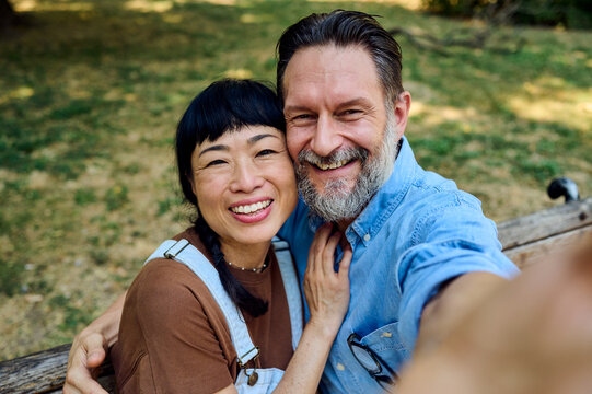 Smiling Mature Mixed Couple Taking Selfie on Park Bench &mdash; Happy Asian Woman and Bearded Man