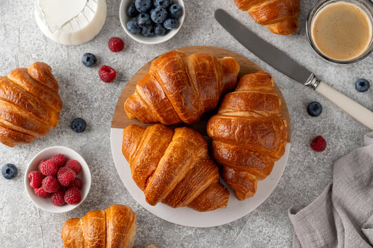 Fresh golden homemade croissants for breakfast with fresh berries, coffee and milk on a board on a gray concrete background. Top view.