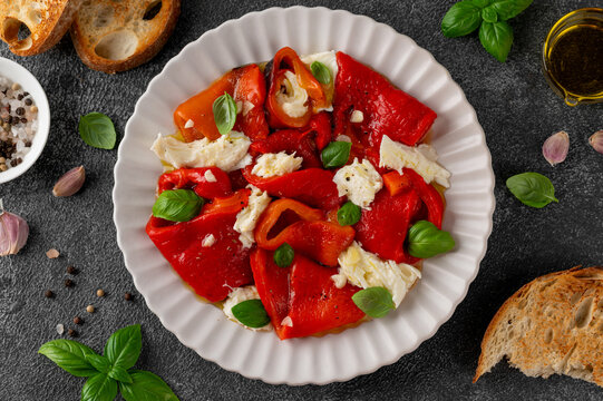 Salad with roasted red peppers and mozzarella in olive oil and garlic on a plate on dark background. Mediterranean appetizer served with bread. Top view.