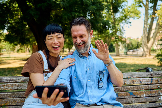 Happy Asian Woman And Bearded Man Video Calling On Smartphone While Sitting On Park Bench