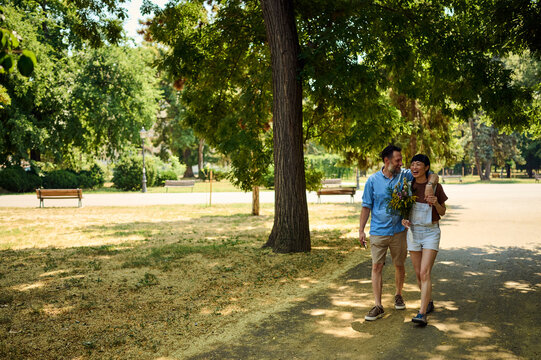 Happy Couple Walking In Sunny City Park Holding Wildflower Bouquet &mdash; Asian Woman And Partner