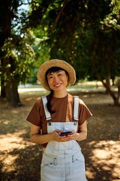 Smiling Asian Woman In Straw Hat And Overalls Using Phone In Sunny Park, Casual Outdoor Lifestyle