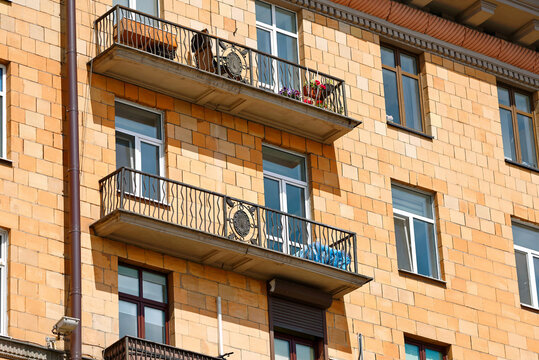 Yellow block masonry building facade, balconies with decorative wrought iron railings glass windows, potted flowers and car tires wrapped in blue plastic storage covers placed on an apartment balcony