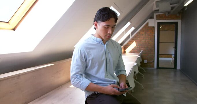 Asian man in mid 20s checking smartphone, tapping and scrolling on long bench beneath skylights