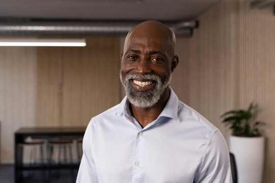 Senior man wearing light button-down shirt smiling and posing in modern office beside white planter