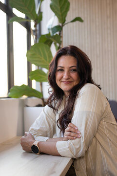 Middle-aged woman leaning on table by window with fig plant, wearing smartwatch and striped top