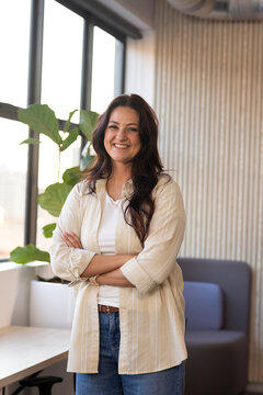 Woman standing arms crossed smiling in modern lounge near windows potted plant wearing jeans