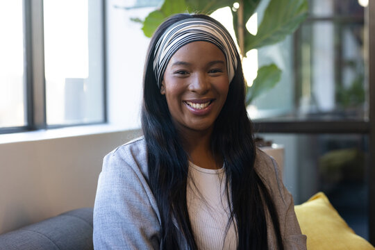 Adult African woman smiling and sitting on gray sofa near large window with plant