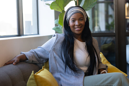 African American woman sitting on gray sofa in modern lounge, wearing blazer, headband, smartwatch